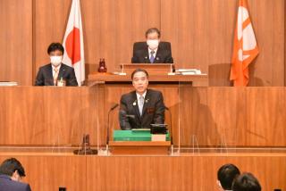 写真：議場で知事議案を説明する知事。後ろに議長と副議長。